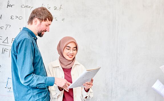 Two people standing in front of a whiteboard with mathematical notations. One holds a piece of paper and appears to be explaining something to the other.